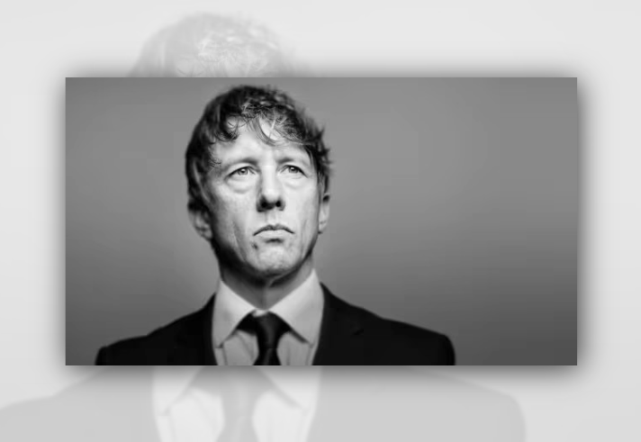 Black-and-white studio portrait of a person in a dark suit jacket, white shirt and dark tie against a plain grey background. The person is shown from the chest up, facing forward with their gaze directed slightly upward. Soft lighting creates contrast across the face and clothing, while the background fades smoothly from darker grey at the edges to lighter grey near the centre.