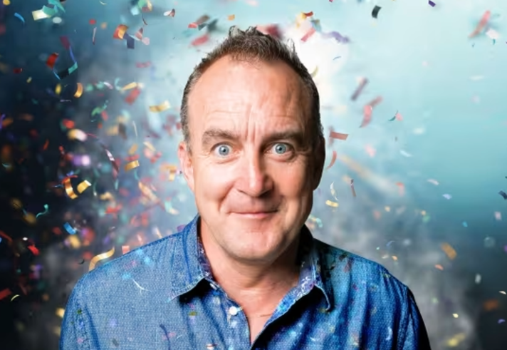 A portrait of Jimeoin. Behind him there is loads of confetti flying about.