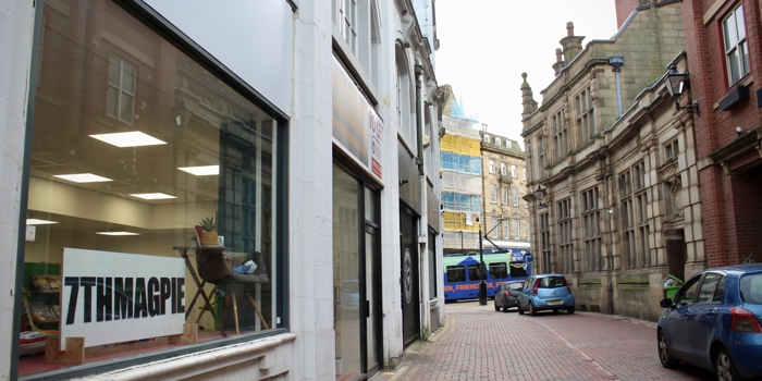 A narrow cobbled street lined with older buildings, with a large shop window in the foreground displaying a sign that reads “7THMAGPIE.” Parked cars and a tram are visible further down the street.