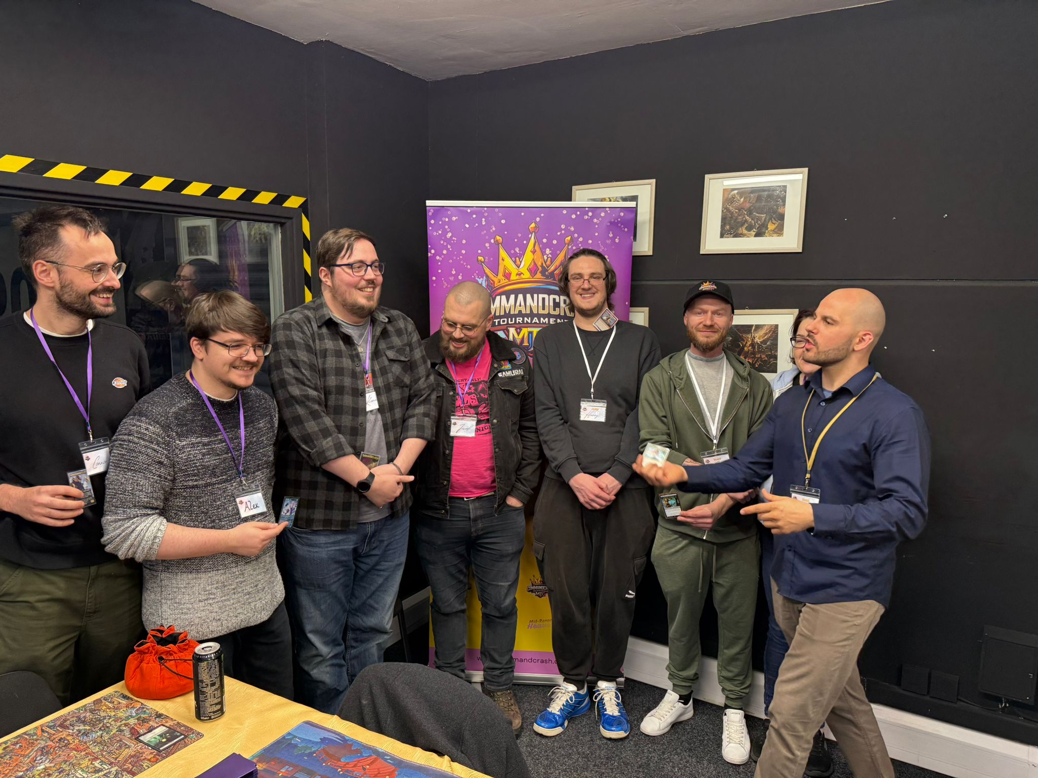  A group of people standing in a gaming room beside a purple CommandCrash tournament banner. A person on the right appears to be awarding or presenting something. Tables in the foreground hold cards, playmats and drinks, creating a lively tournament atmosphere.