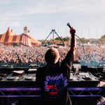 A DJ stands behind a mixing console on an outdoor stage, holding a microphone up toward a large crowd. The audience stretches far into the distance, with a colorful striped circus-style tent and a tall amusement ride visible in the background under a clear blue sky.