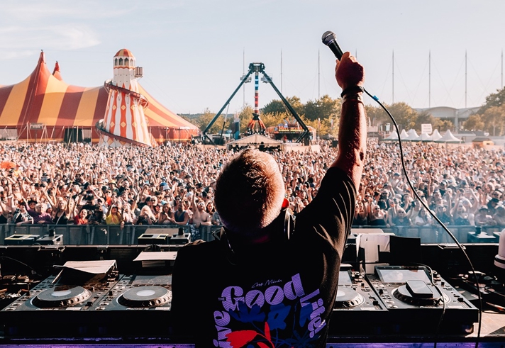 A DJ stands behind a mixing console on an outdoor stage, holding a microphone up toward a large crowd. The audience stretches far into the distance, with a colorful striped circus-style tent and a tall amusement ride visible in the background under a clear blue sky.