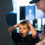 A young boy plays an arcade game, staring wide-eyed at the screen with his mouth open in amazement and both hands on his head. Beside him, an adult wearing a black cap smiles and watches the action. The arcade cabinet fills the left side of the image, while softly blurred gaming machines and lights in the background create a lively, family-friendly gaming environment.