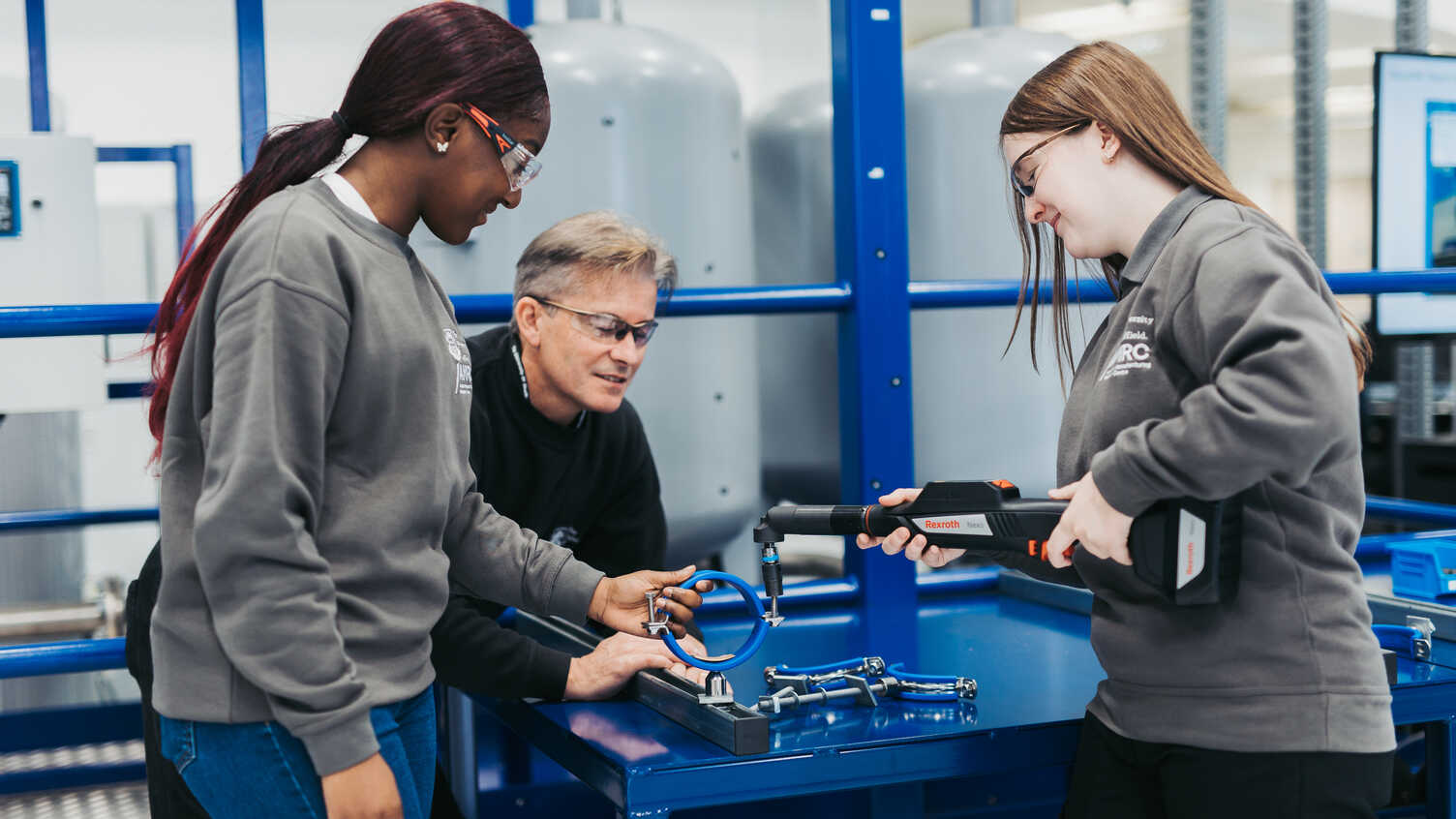 Three individuals working together in a technical workshop setting. They are using a handheld scanning device on a blue circular metal component placed on a workbench, surrounded by other metal parts and fixtures. The background includes large industrial equipment and blue structural frames.