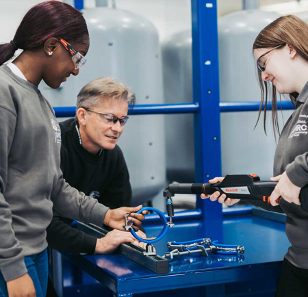 Three individuals working together in a technical workshop setting. They are using a handheld scanning device on a blue circular metal component placed on a workbench, surrounded by other metal parts and fixtures. The background includes large industrial equipment and blue structural frames.