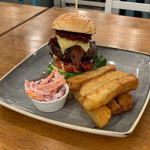 A tall burger , a stack of chunky square cut chips and a bowl of coleslaw on a square plate.