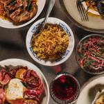 A tabletop spread of vibrant dishes including grilled meat in sauce, a bowl of spiced rice topped with crispy onions, a salad with red leaves and herbs, and a plate with creamy cheese and roasted vegetables, alongside a glass of red drink.