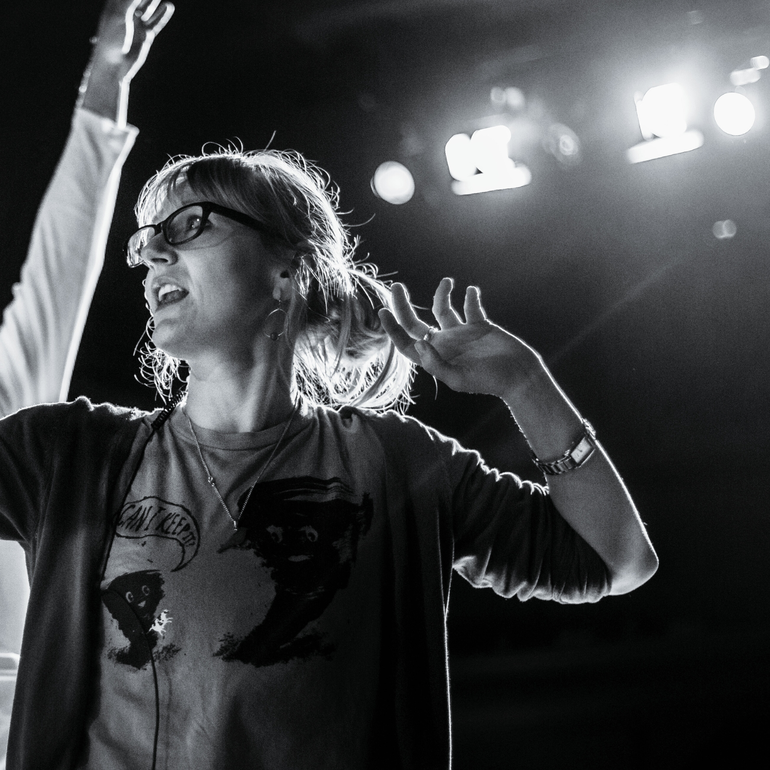 A black and white photo standing under stage lights, with one arm slightly raised. Another person's arm is just coming into shot.