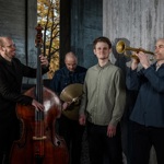 A four‑piece jazz group stands in a concrete architectural space. One musician plays a double bass, another holds cymbals, a third stands in the centre, and the fourth plays a trumpet. Sunlight filters through an opening behind them, creating contrast between the warm outdoor light and the cool, shadowed interior.