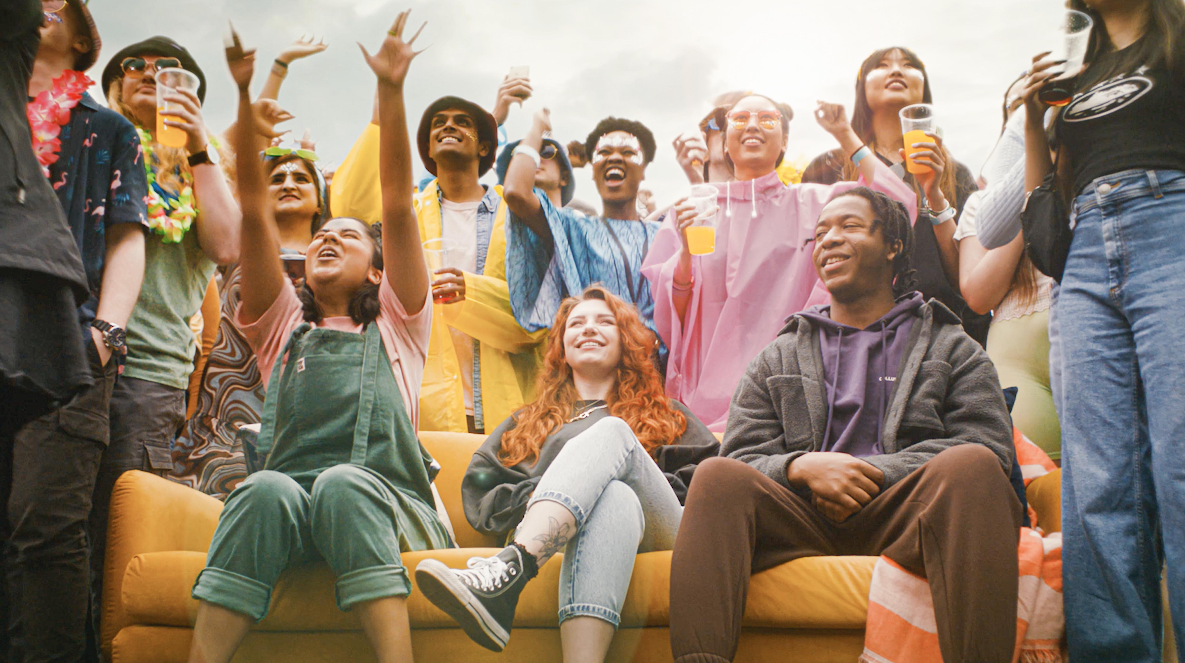 A lively outdoor gathering with a large group of people standing and sitting around a bright yellow couch. Many are wearing colorful clothing and accessories like leis and ponchos, holding drinks, and raising their arms in celebration. The background shows an overcast sky, suggesting a festival or party atmosphere.