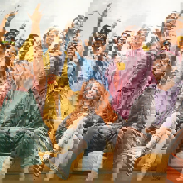 A lively outdoor gathering with a large group of people standing and sitting around a bright yellow couch. Many are wearing colorful clothing and accessories like leis and ponchos, holding drinks, and raising their arms in celebration. The background shows an overcast sky, suggesting a festival or party atmosphere.