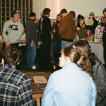 Visitors browse stalls and socialise at a busy indoor arts and craft market. Tables display handmade goods, prints and jewellery, while groups of attendees stand chatting around the venue. In the foreground, people sit at a wooden table, and in the background market traders showcase products beneath warm lighting. Glass-block walls and an industrial interior create a relaxed, community-focused atmosphere.
