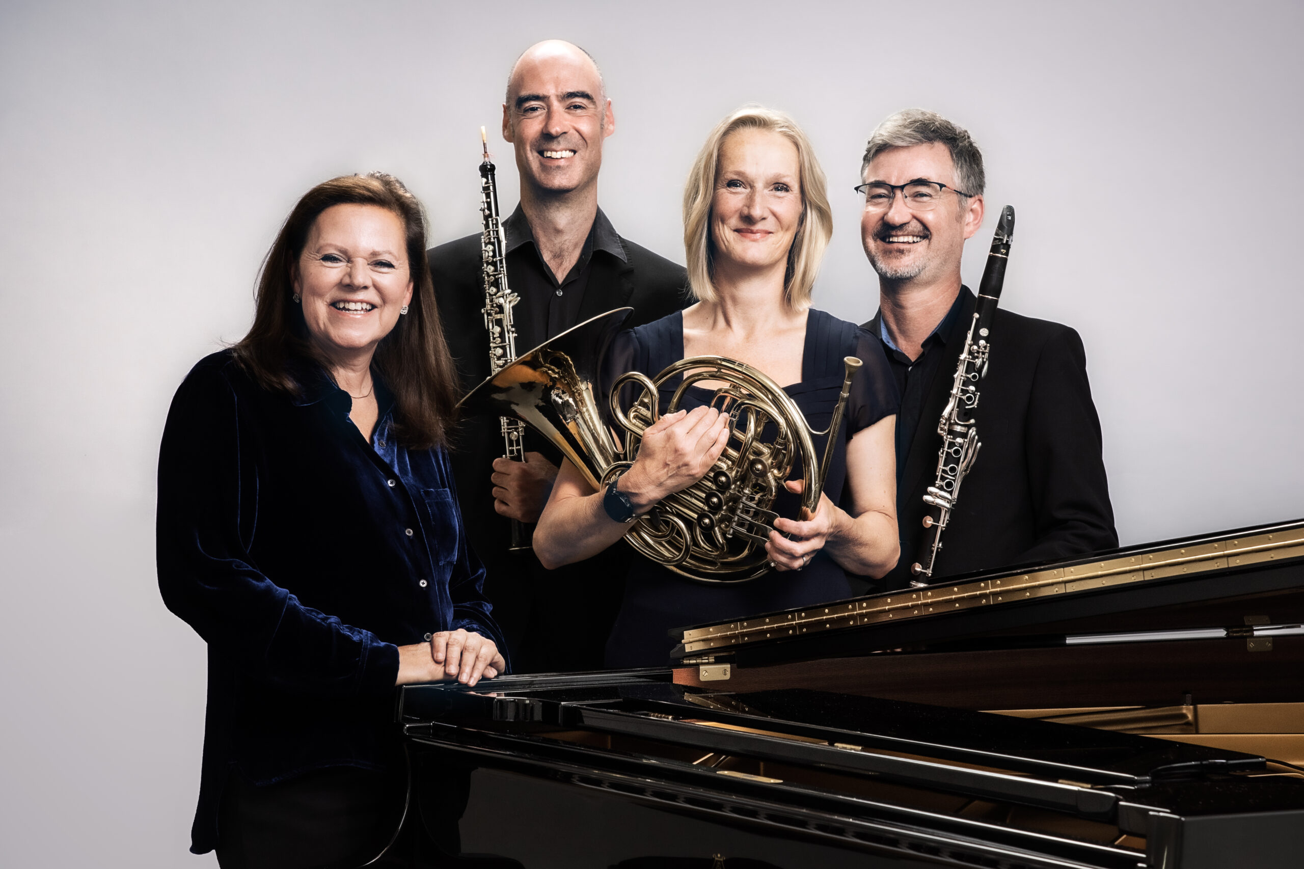 Kathryn Stott and members of Ensemble 360 (who are holding their musical instruments) stand behind a grand piano, smiling at the camera.