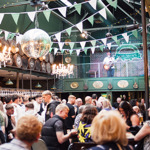 Busy indoor venue with a crowd gathered beneath green bunting, string lights, and chandeliers. Barrels line the upper wall, and a mirrored disco ball hangs overhead. People stand closely together around tables and the bar. Above, a person plays guitar on a balcony stage against a brick wall with a venue sign. The space has a lively, decorated pub or event atmosphere.