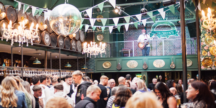 Busy indoor venue with a crowd gathered beneath green bunting, string lights, and chandeliers. Barrels line the upper wall, and a mirrored disco ball hangs overhead. People stand closely together around tables and the bar. Above, a person plays guitar on a balcony stage against a brick wall with a venue sign. The space has a lively, decorated pub or event atmosphere.