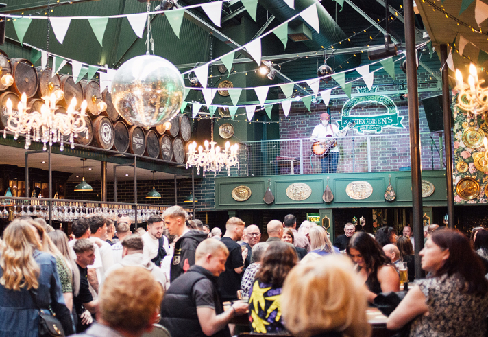 Busy indoor venue with a crowd gathered beneath green bunting, string lights, and chandeliers. Barrels line the upper wall, and a mirrored disco ball hangs overhead. People stand closely together around tables and the bar. Above, a person plays guitar on a balcony stage against a brick wall with a venue sign. The space has a lively, decorated pub or event atmosphere.