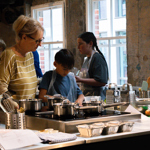 People take part in a cooking workshop in a modern kitchen classroom. Several participants wearing aprons stand around workstations with saucepans, mixing bowls, whisks, graters and utensils. An adult assists a child preparing ingredients at the centre, while others work nearby. Chopped vegetables, herbs and cooking trays are arranged on the counters. Large windows and exposed concrete walls create a bright, industrial-style setting.