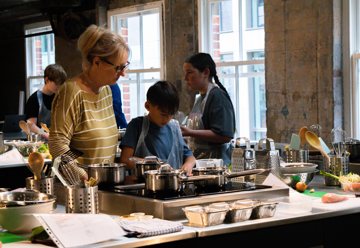 People take part in a cooking workshop in a modern kitchen classroom. Several participants wearing aprons stand around workstations with saucepans, mixing bowls, whisks, graters and utensils. An adult assists a child preparing ingredients at the centre, while others work nearby. Chopped vegetables, herbs and cooking trays are arranged on the counters. Large windows and exposed concrete walls create a bright, industrial-style setting.
