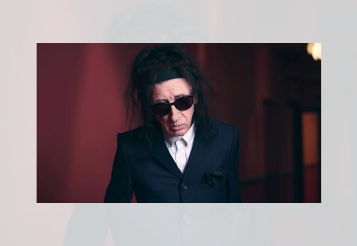 Studio-style portrait of a person with long dark hair wearing dark sunglasses, a dark tailored suit jacket and a white collared shirt. The person stands indoors against a softly lit background in deep red and burgundy tones. The image is closely cropped from the waist up, with warm lighting creating contrast between the dark clothing and the rich-coloured surroundings.