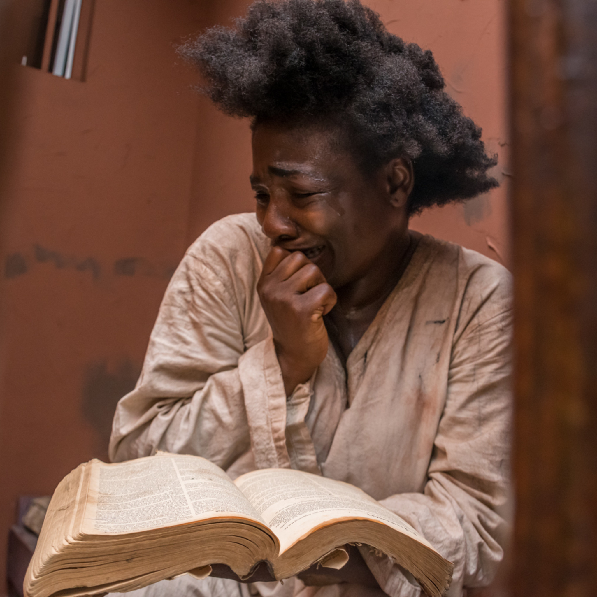 A person in loose, worn clothing sits beside a large open book with aged pages. The person rests one hand against their mouth while looking down at the book. The scene is framed by reddish-brown walls and lit with warm, soft light. The thick volume dominates the foreground, with visible columns of text on its pages, creating a contemplative indoor setting.