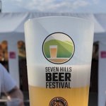 A reusable cup filled with pale beer held up in front of a white marquee tent. The cup displays the Seven Hills Beer Festival logo featuring stylized hills and a pint glass.