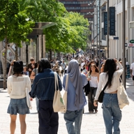 Four people walking down a busy pedestrian shopping street lined with stores and outdoor seating. Visible shop signs include “Caffè Nero” on the left and “Primark” on the right. The street is shaded by green trees, and the background shows a large crowd of people, modern buildings, and bright daylight.