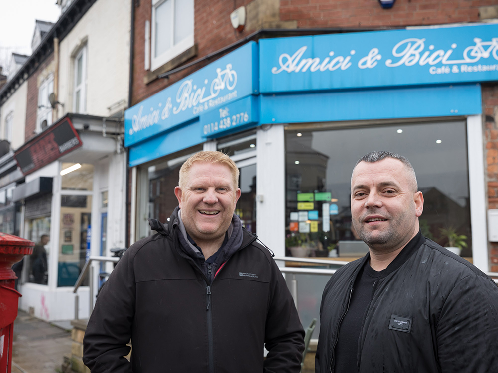 Two men, standing outside a shop called Amici & Bici, are looking at the camera and smiling.