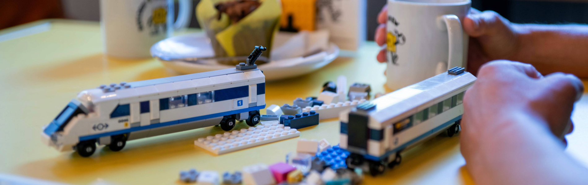 A wooden table in a cafe. On the table are two hot drinks and a partially built Lego© train, along with the instructions and the remaining pieces.