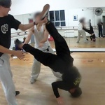 People practise breakdancing in a mirrored dance studio with wooden floors and ballet barres along the walls. In the foreground, one dancer balances upside down with legs raised while two others support the move. Reflections in the wall-length mirror show additional participants rehearsing similar techniques. The bright studio includes windows, a wall-mounted fan and exercise equipment, capturing a dance training session focused on acrobatic floorwork.