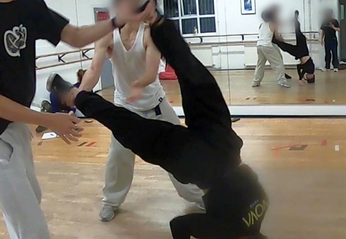 People practise breakdancing in a mirrored dance studio with wooden floors and ballet barres along the walls. In the foreground, one dancer balances upside down with legs raised while two others support the move. Reflections in the wall-length mirror show additional participants rehearsing similar techniques. The bright studio includes windows, a wall-mounted fan and exercise equipment, capturing a dance training session focused on acrobatic floorwork.