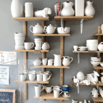 Shelves displaying unpainted ceramic pottery, including mugs, teapots, vases, bowls, and figurines, inside a ceramics painting studio.