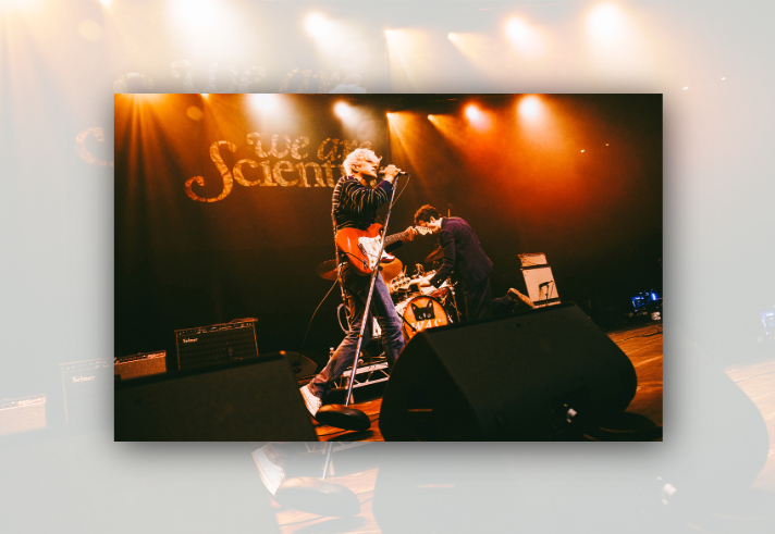 Live performance by rock band We Are Scientists on a theatre stage. A guitarist and vocalist with a red electric guitar performs at a microphone under orange stage lights, while another musician kneels beside equipment near the drum kit. Large black stage monitors fill the foreground, amplifiers sit at the sides, and the band's name, “We Are Scientists”, is displayed in large lettering behind the performers.