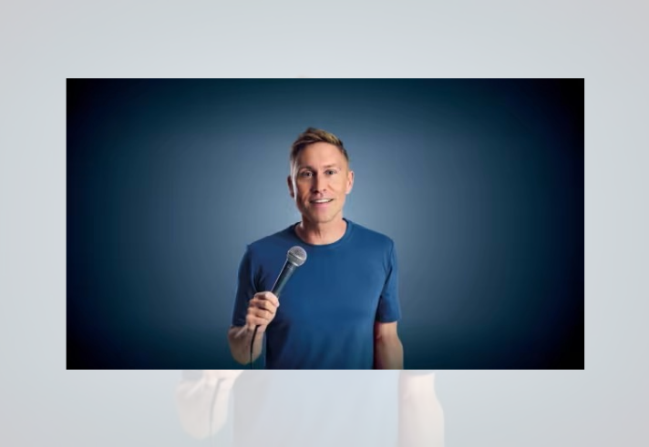 Promotional-style portrait of a person standing against a blue gradient background, holding a microphone in one hand and facing the camera. The person wears a plain blue T-shirt and is centred in the frame. Studio lighting highlights the figure, with a dark blue vignette around the edges creating a simple, professional backdrop.