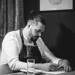 A black and white picture of a man in chef's clothing, writing at a tabel.