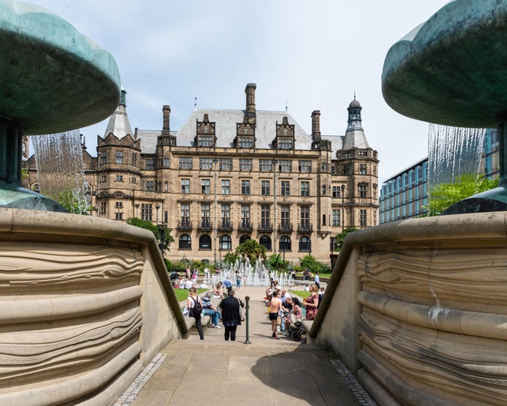 Two fountains in the Peace Gardens frame Sheffield Town Hall in the city centre. The Peace Gardens is full of people enjoying the sunshine.