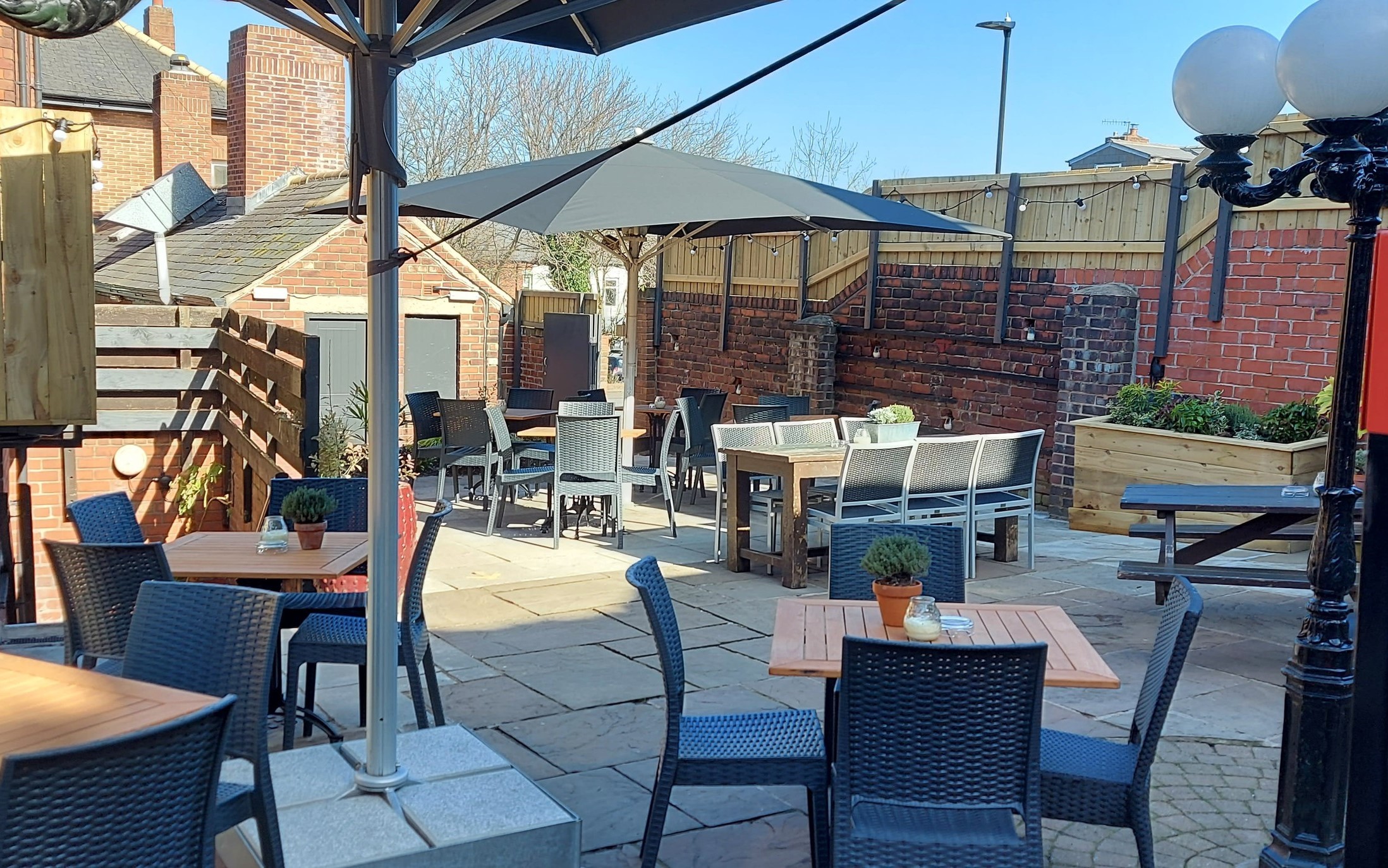 An outdoor pub terrace with wicker chairs, wooden tables and large parasols, surrounded by brick walls and planters on a sunny day.