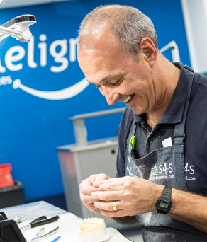 A smiling male technician, in work-wear and an apron, sits working on a set of hi-tech braces for the dental sector.