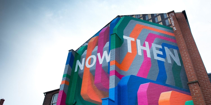 Colorful geometric mural on the side of a tall brick building featuring bold overlapping shapes in orange, pink, green, blue, and gray, with large white text reading ‘NOW THEN’ against a bright sky.