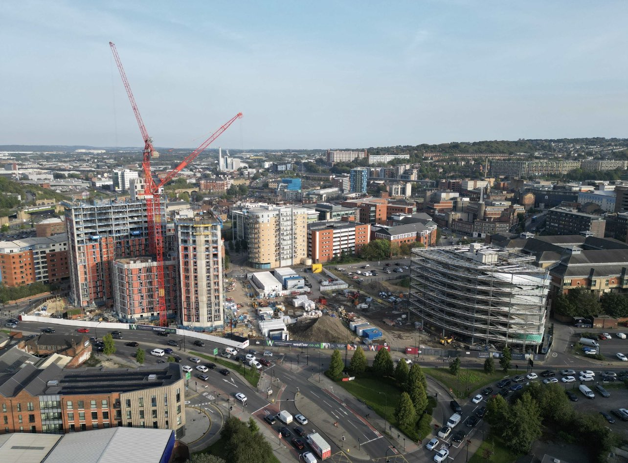 Aerial view of a large urban construction site surrounded by modern buildings. Two tall red cranes dominate the scene, working on high-rise structures with exposed concrete and steel. Nearby, a circular building is covered in scaffolding, and several mid-rise apartment blocks are visible. The foreground shows a busy roundabout with cars, while the background extends to a hilly cityscape under a clear sky.