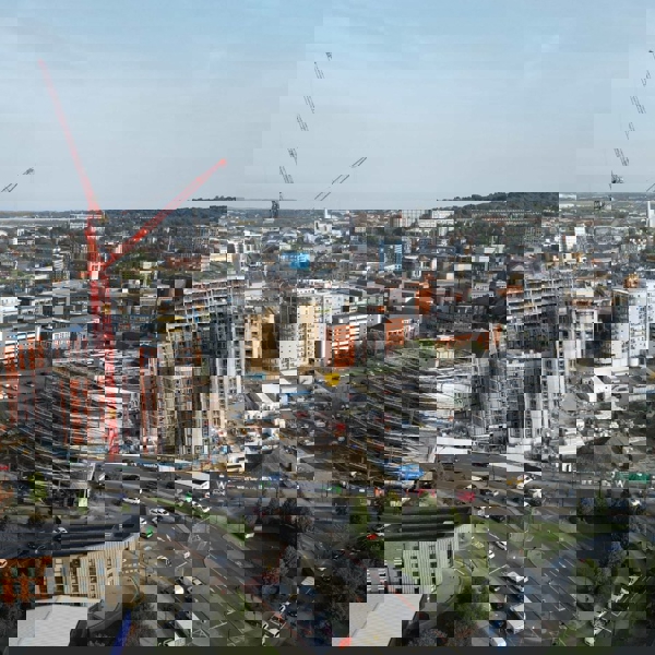 Aerial view of a large urban construction site surrounded by modern buildings. Two tall red cranes dominate the scene, working on high-rise structures with exposed concrete and steel. Nearby, a circular building is covered in scaffolding, and several mid-rise apartment blocks are visible. The foreground shows a busy roundabout with cars, while the background extends to a hilly cityscape under a clear sky.