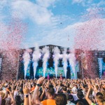 A packed audience at an outdoor music festival cheers in front of the “90’s Fest Summer Bowl” stage as large bursts of white smoke and red confetti erupt into the sky. The stage displays vibrant visuals, and the atmosphere is festive and colorful.