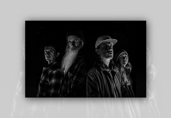 Black-and-white portrait of four people standing closely together against a dark brick wall. Dramatic side lighting illuminates their faces and clothing while much of the background remains in shadow. One person has a long white beard and a knit hat, another wears a baseball cap and jacket, and the others wear casual outdoor clothing. The high-contrast monochrome style creates a striking, atmospheric group portrait.