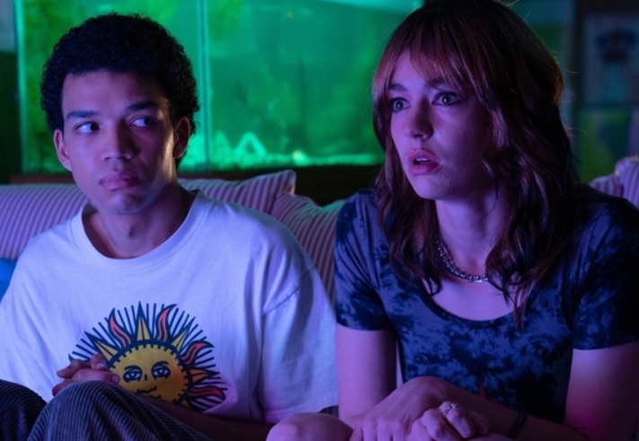 A still from the film "I Saw The TV Glow": two young people are sat on a sofa in a dimly lit room.