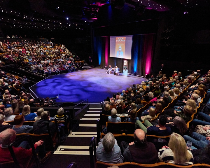 A packed theatre with tiered seating surrounding a central stage where two people are seated for a live talk under colourful lighting.