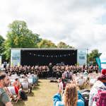 Outdoor event with a large stage featuring a choir performance, rows of deck chairs filled with spectators, and colourful bunting overhead. Inflatable structures and trees surround the area under a partly cloudy sky.