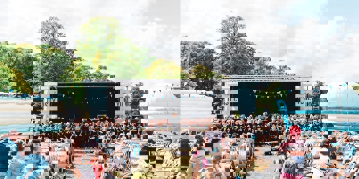 Outdoor event with a large stage featuring a choir performance, rows of deck chairs filled with spectators, and colourful bunting overhead. Inflatable structures and trees surround the area under a partly cloudy sky.