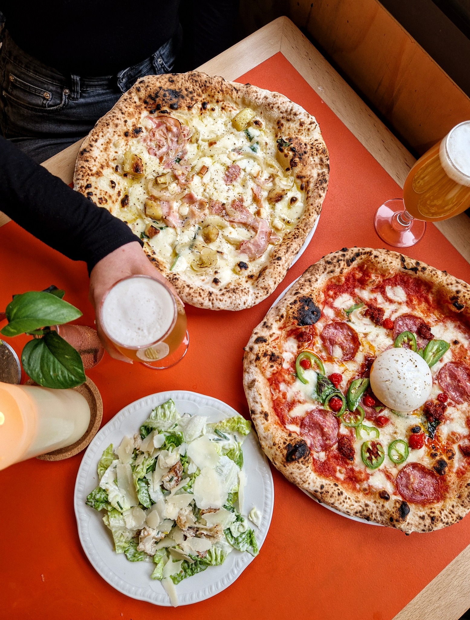 Two Neapolitan pizzas on a table, one topped with sausage and potatoes and the other with pepperoni and jalapeños, served with a Caesar salad and two glasses of beer.