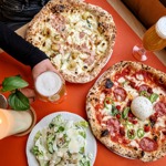 Two Neapolitan pizzas on a table, one topped with sausage and potatoes and the other with pepperoni and jalapeños, served with a Caesar salad and two glasses of beer.