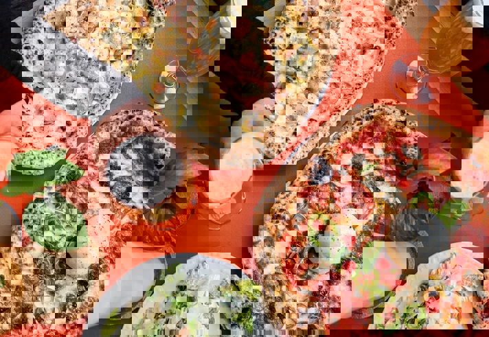 Two Neapolitan pizzas on a table, one topped with sausage and potatoes and the other with pepperoni and jalapeños, served with a Caesar salad and two glasses of beer.