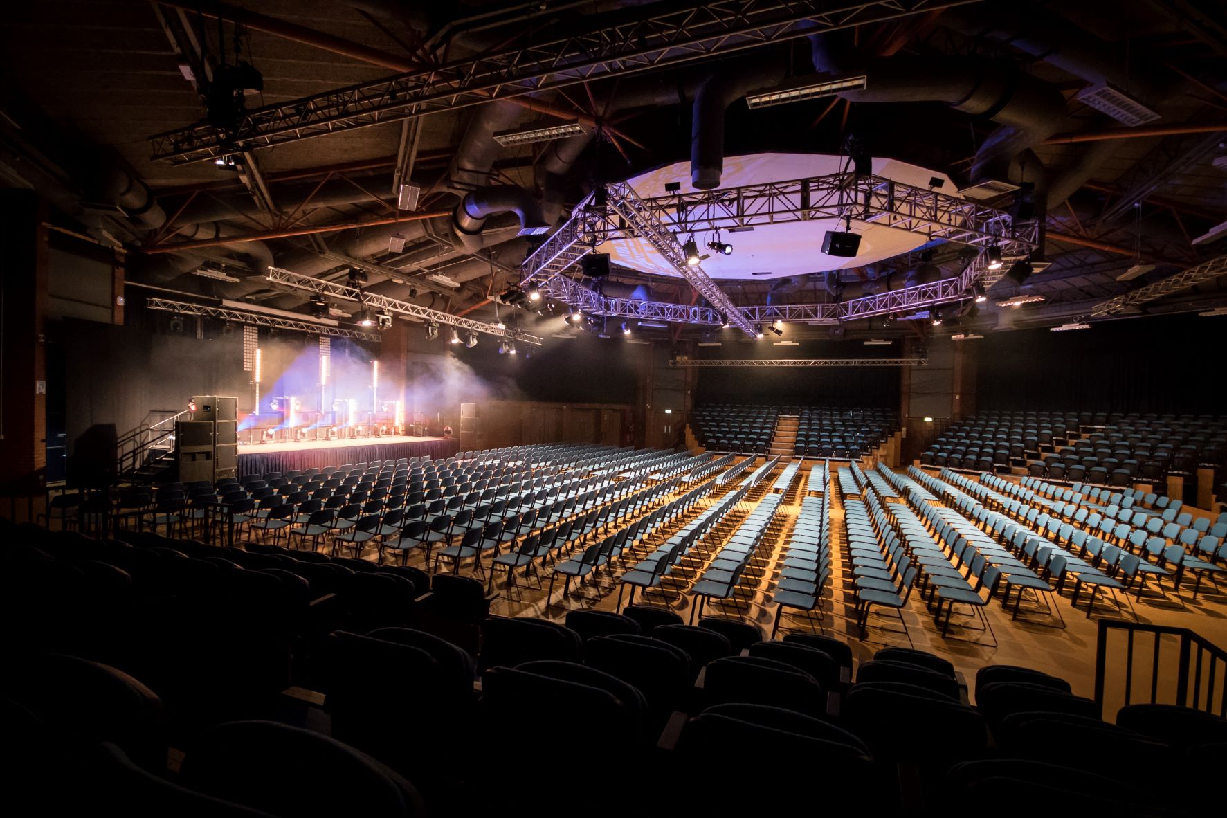 Large auditorium set up in theatre style with rows of blue chairs facing a stage. The stage is lit with spotlights and has a podium and equipment, while the ceiling features exposed beams, ducts, and a circular truss with lighting rigs. The space is dimly lit except for the illuminated stage area.